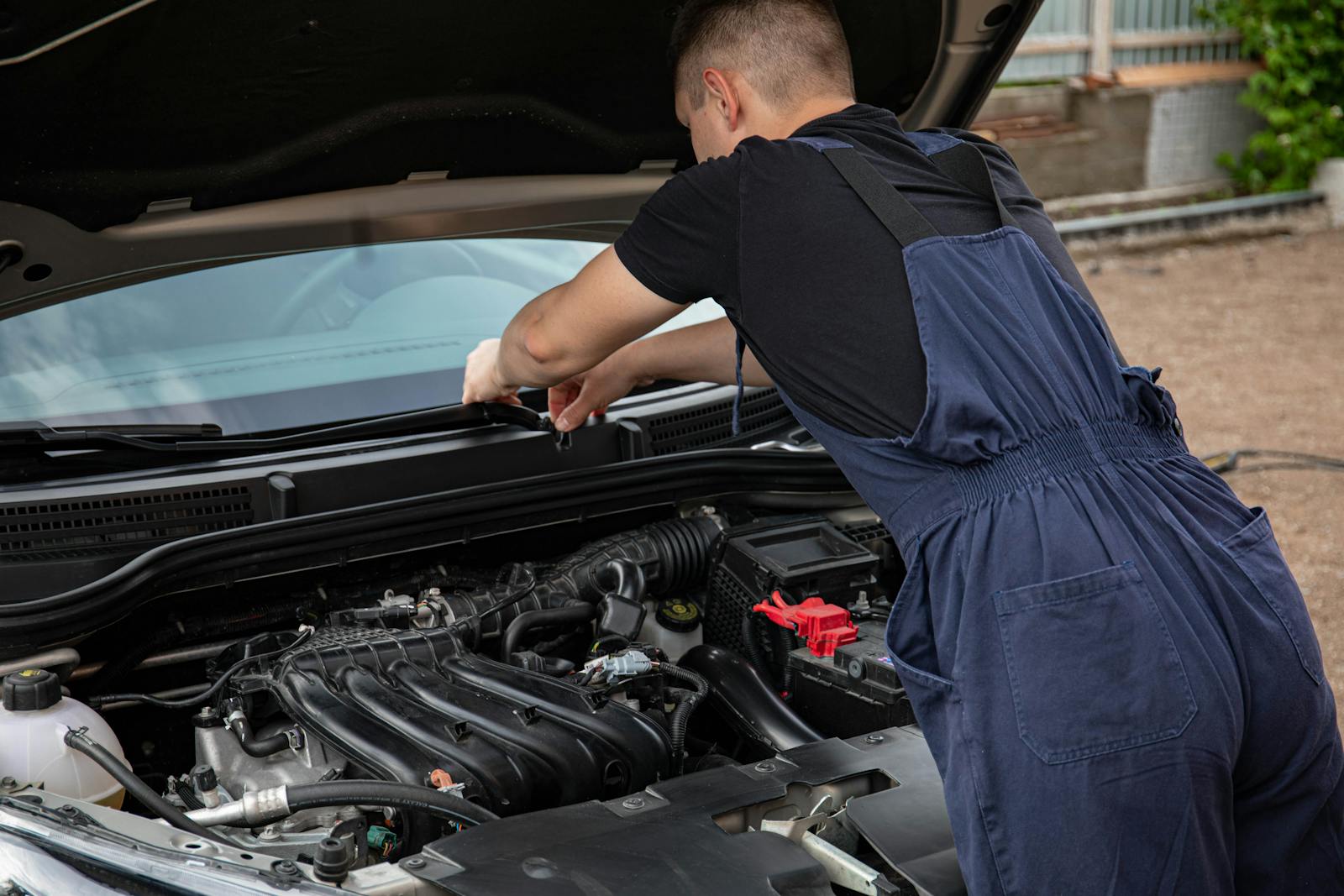 A mechanic in blue overalls works on a car engine outdoors, showcasing vehicle maintenance skills.