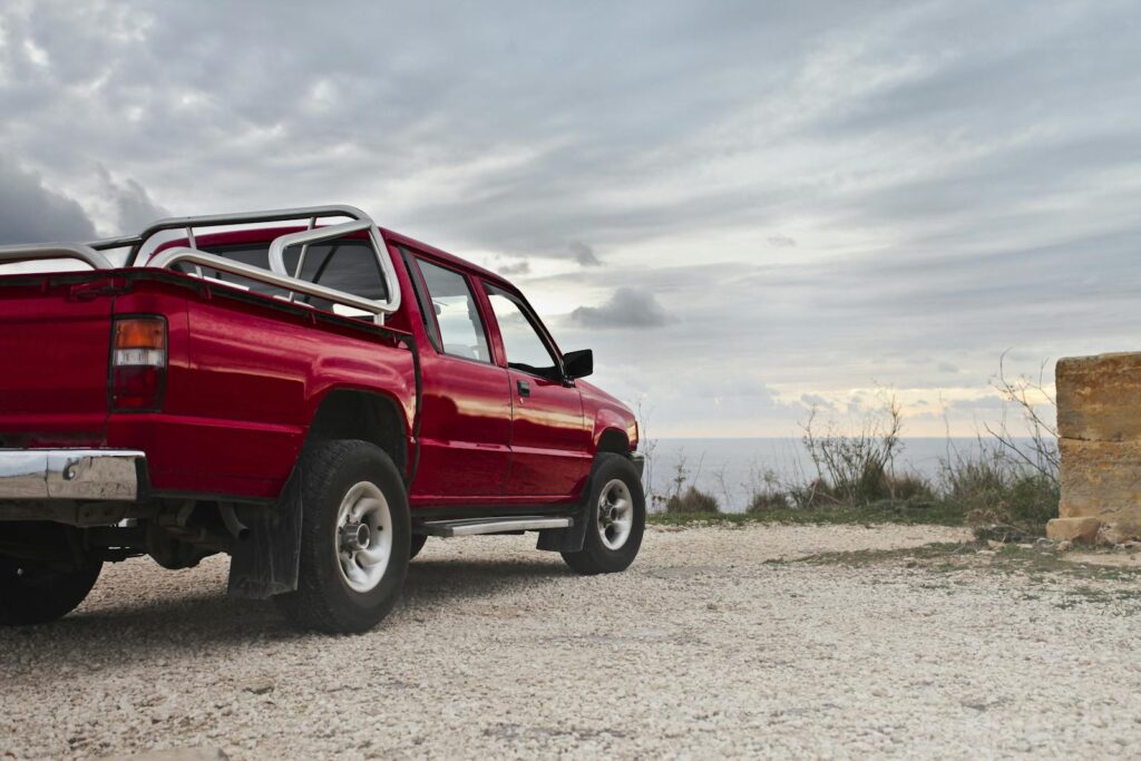 A red pickup truck parked by the rocky coastline under a cloudy sky in Mgarr, Malta.