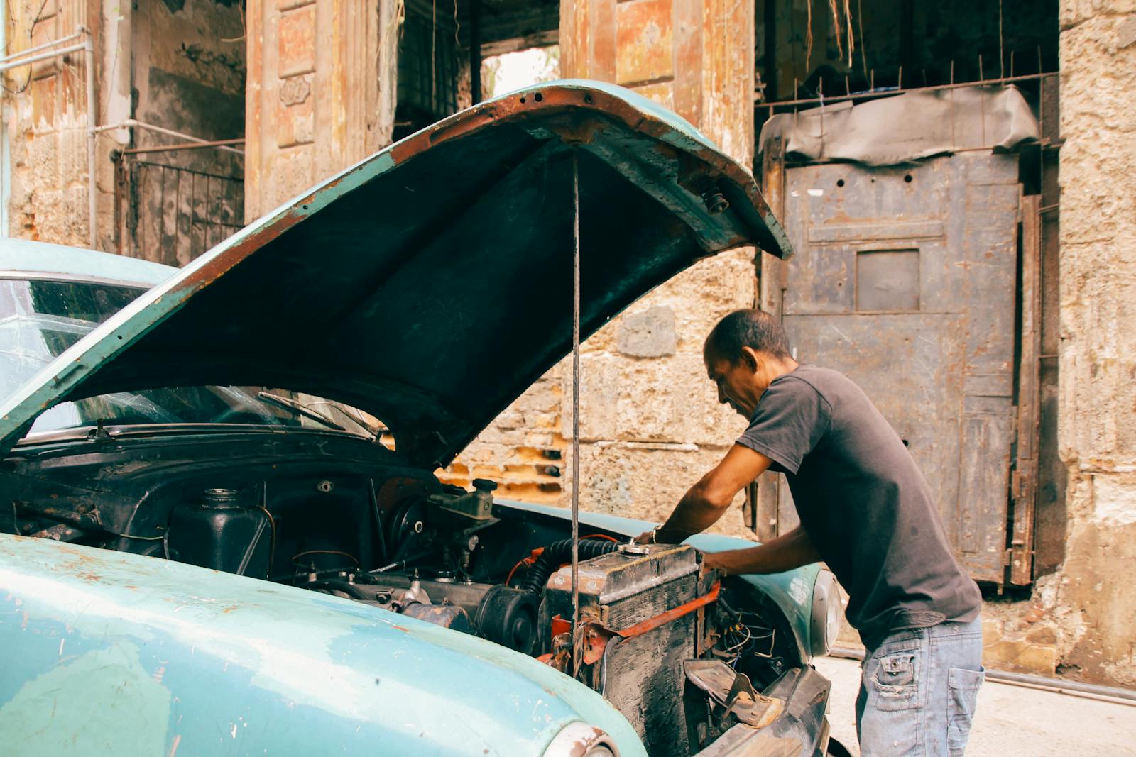 A man working on an old vintage car engine in a rustic outdoor garage setting.