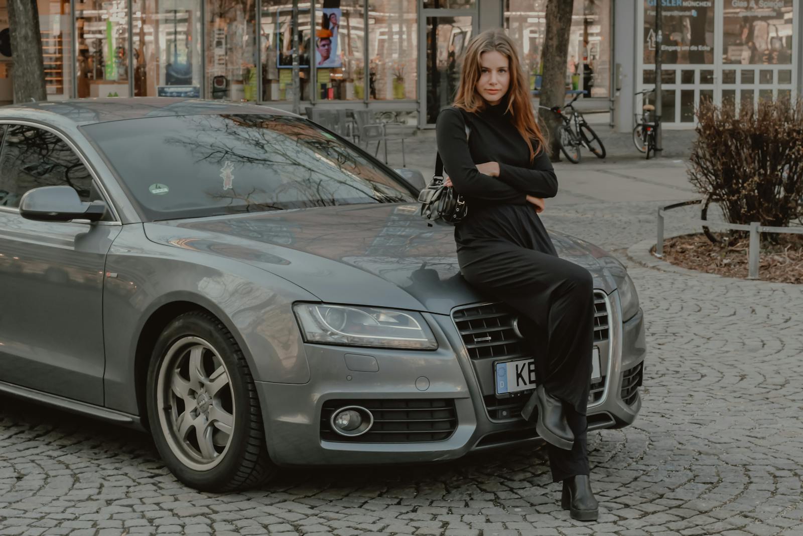 Stylish woman in a black outfit poses on a luxury car in a cityscape with cobblestone streets.