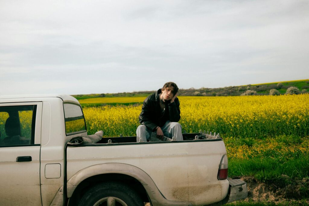 Young man in a leather jacket sitting in a pickup truck amidst a vibrant yellow field.