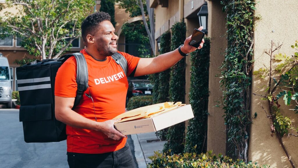 Smiling deliveryman with smartphone and package outside a house.