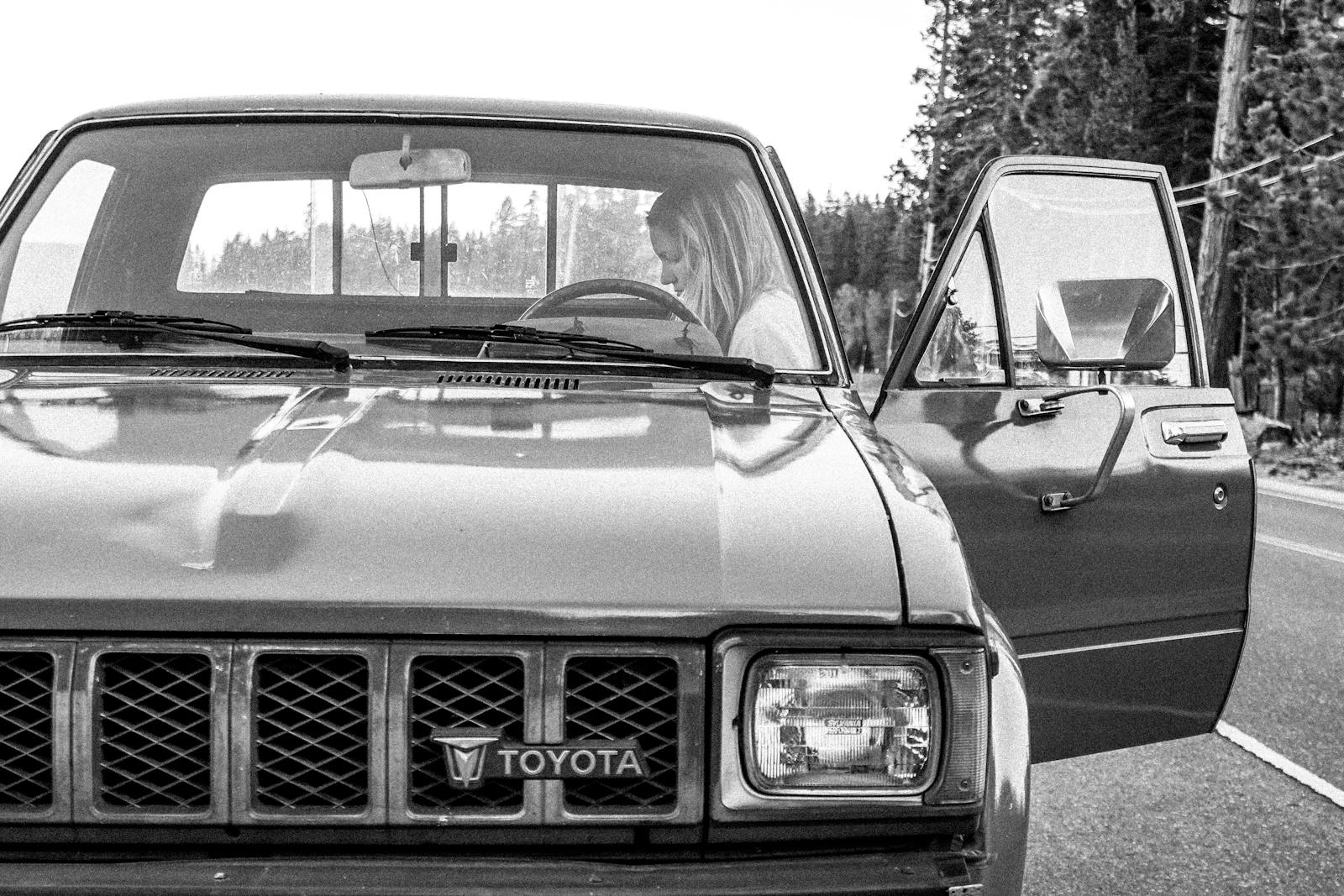 Black and white image of a woman in a vintage Toyota truck parked on a scenic road.