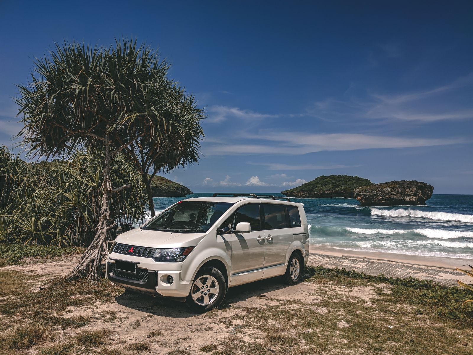 A white SUV parked on a scenic beach in Pacitan, Indonesia, with clear blue sky and ocean waves.