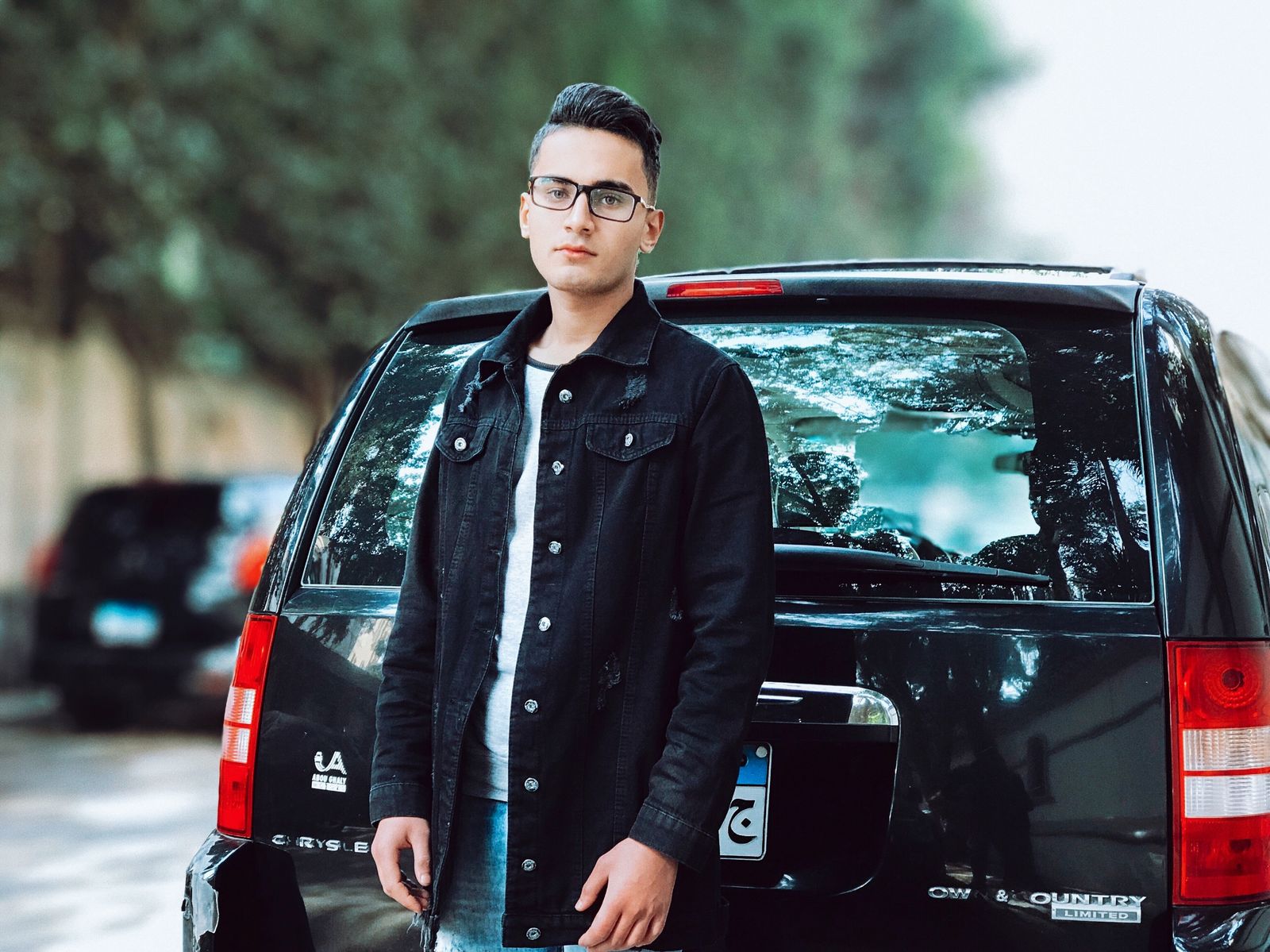 Young man in denim jacket posing confidently with a black SUV in an outdoor setting. Ideal for lifestyle and fashion concepts.
