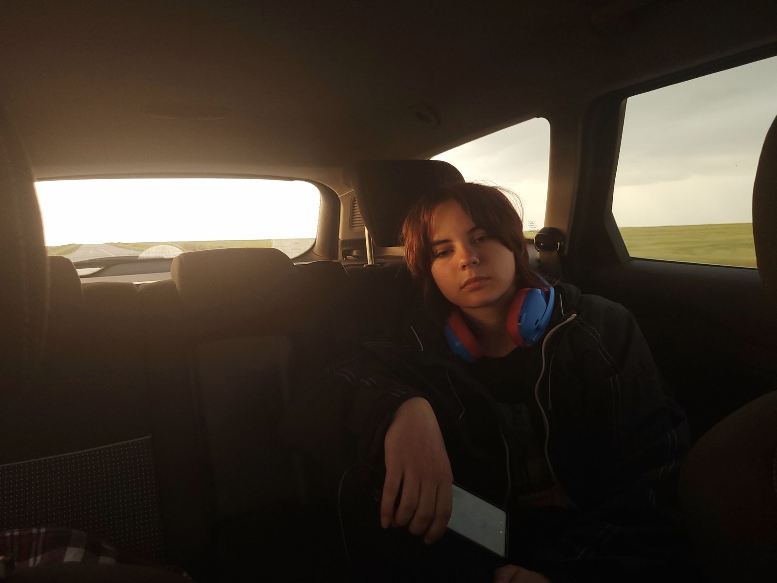 Teenage girl sitting in car, relaxing with headphones during sunset drive.