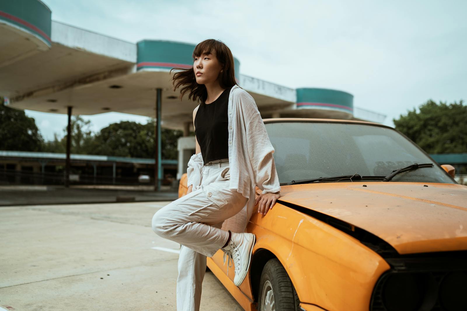 Slender young Asian female in casual wear looking away while standing leaned on one leg near old fashioned car parked on asphalt road on windy weather under cloudy sky