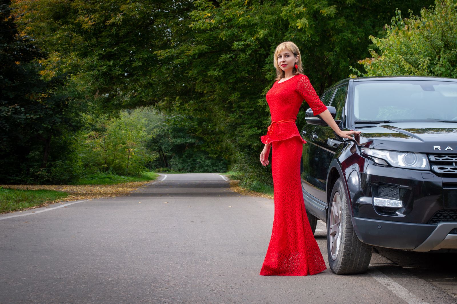 Elegant woman in a striking red dress posing by a black SUV on a secluded road, surrounded by lush greenery.