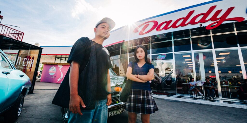 Two young people stand outside a building with "daddy" sign.