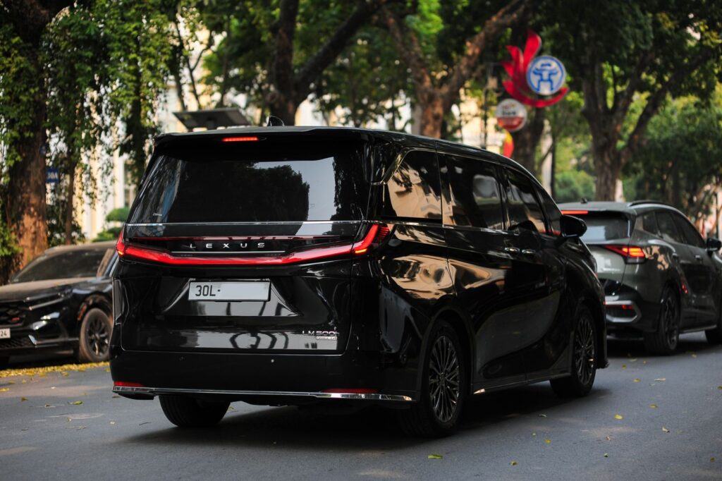 Black Lexus van driving along a tree-lined street in an urban area during daylight.