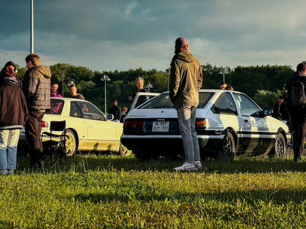 Enthusiasts gathered around two vintage cars on grass.