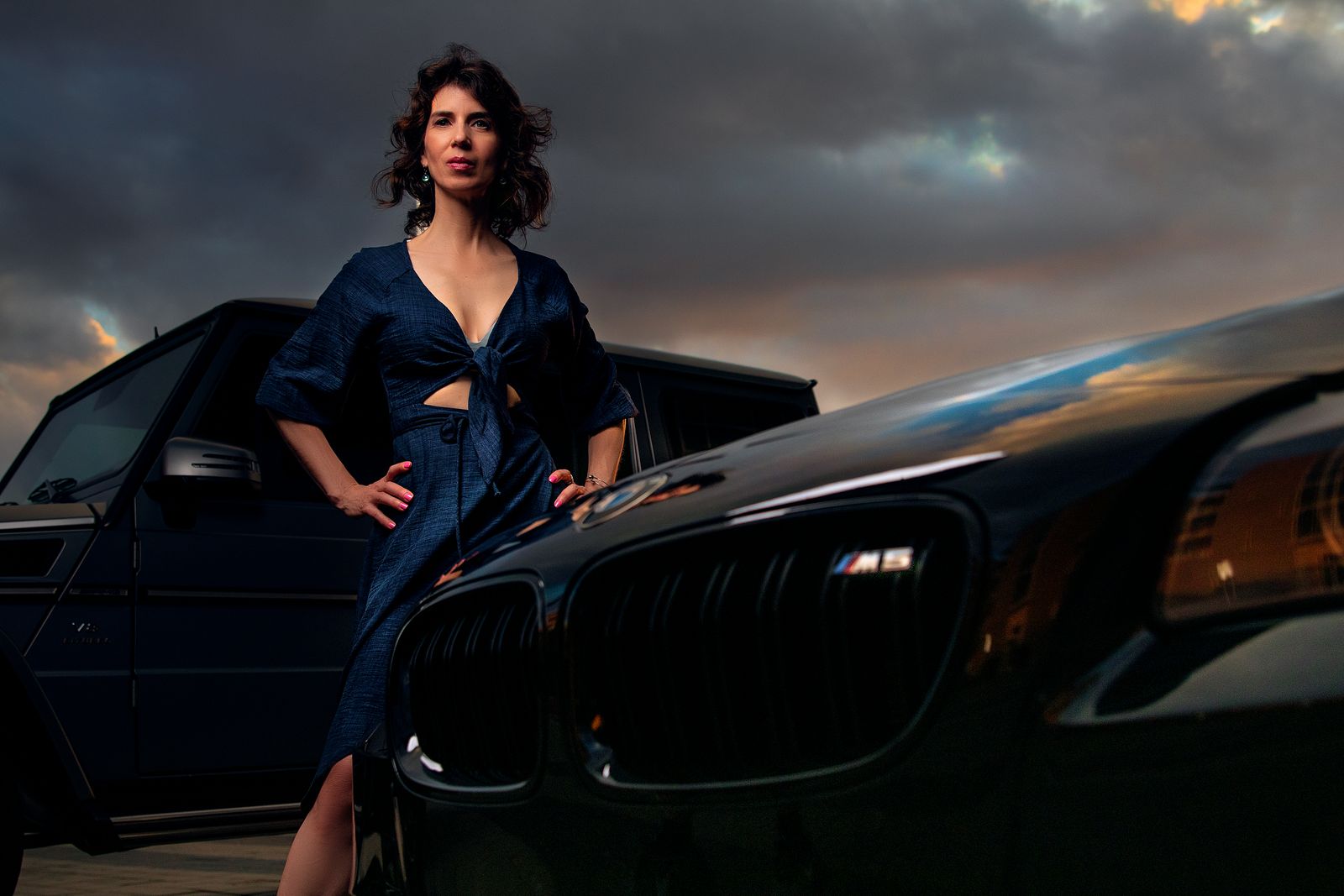 Elegant woman stands confidently between luxury vehicles at sunset.