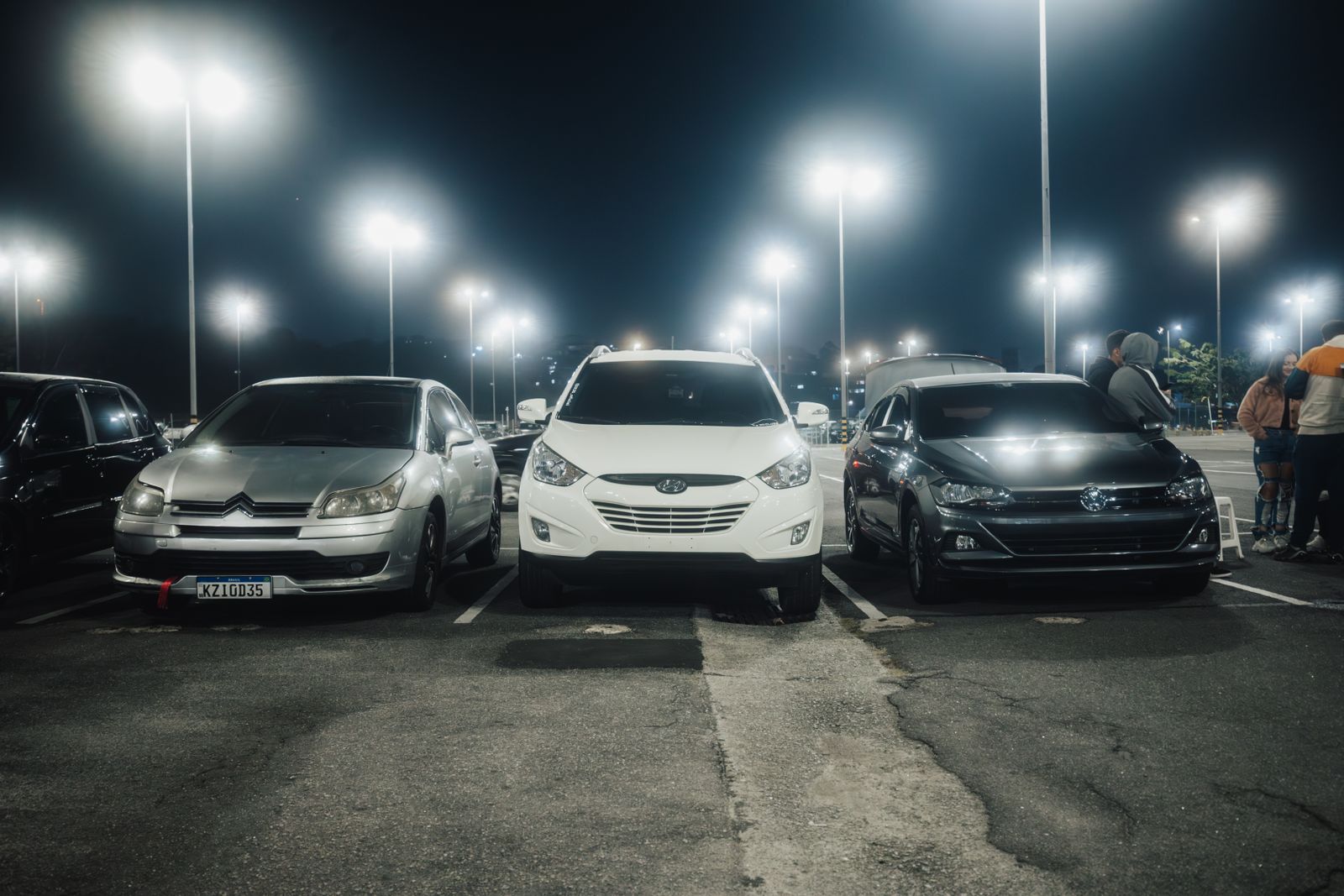 Night scene in a well-lit parking lot featuring various parked cars and street lamps.