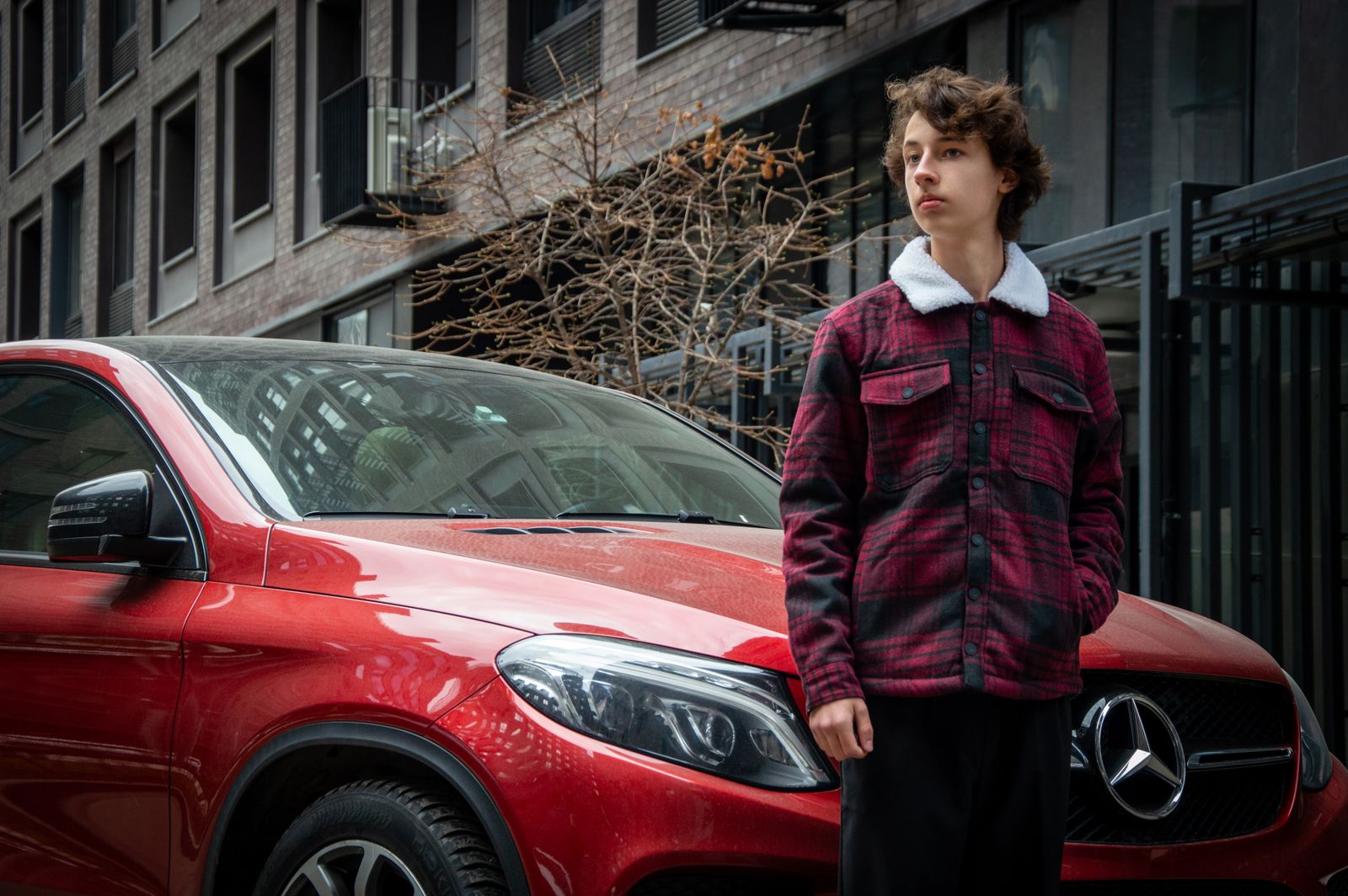 Young adult in a plaid jacket stands confidently by a luxury red car in an urban environment.