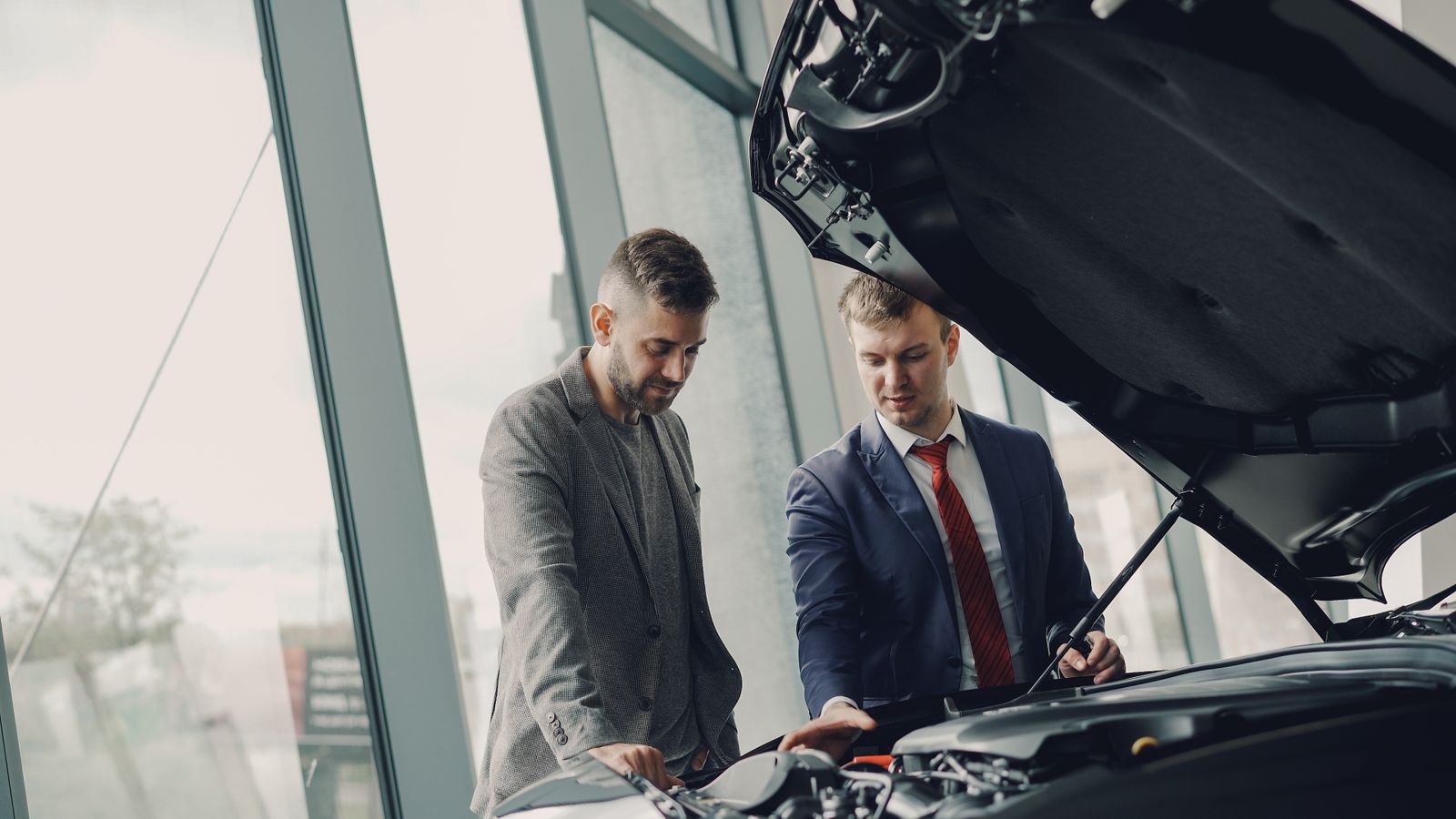 Two men inspecting a car's engine hood in a dealership showroom.