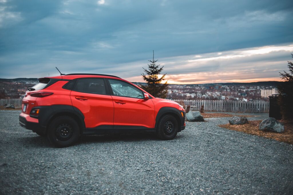 Modern red car parked on shabby road under evening sky full of clouds over cityscape