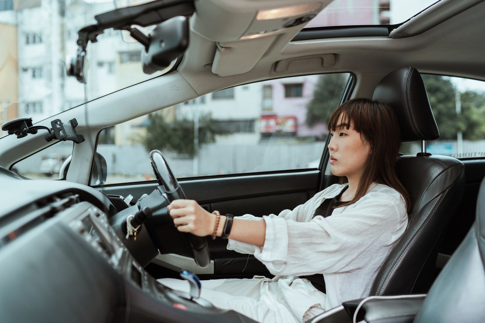 Side view unemotional Asian female in casual white clothes steering right hand drive car along urban streets in daytime