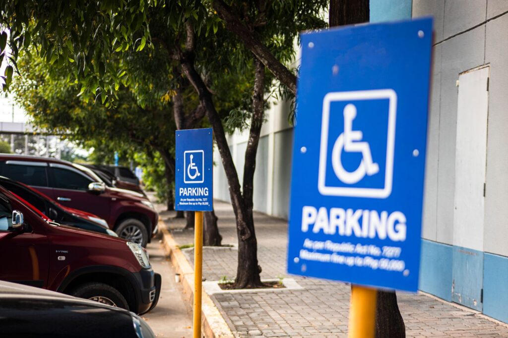 Disabled sign hanging on post on parking lot with row of cars under tree branches