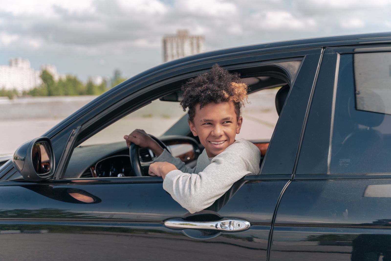 Teenager with curly hair smiling while sitting in car outdoors on a sunny day.