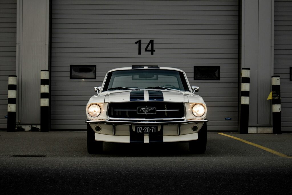 A white Ford Mustang with blue racing stripes parked before a garage door