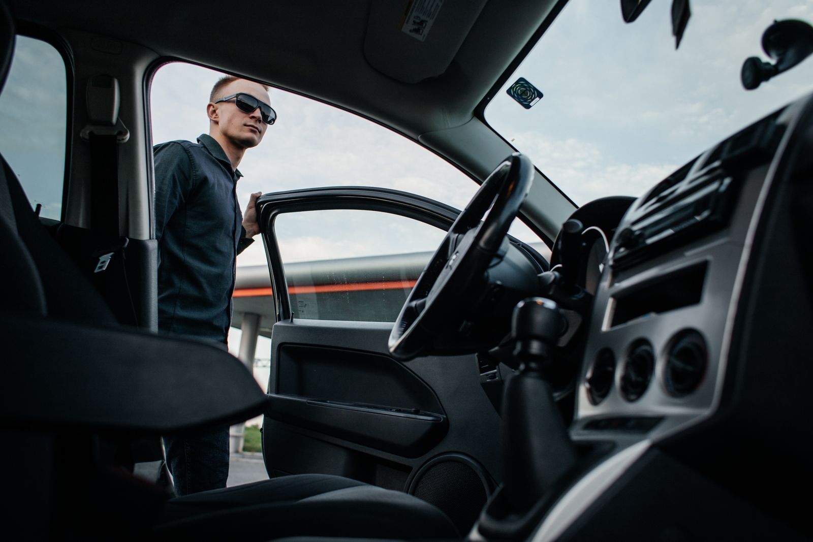 A man in sunglasses stands by an open car door, viewed from the interior.