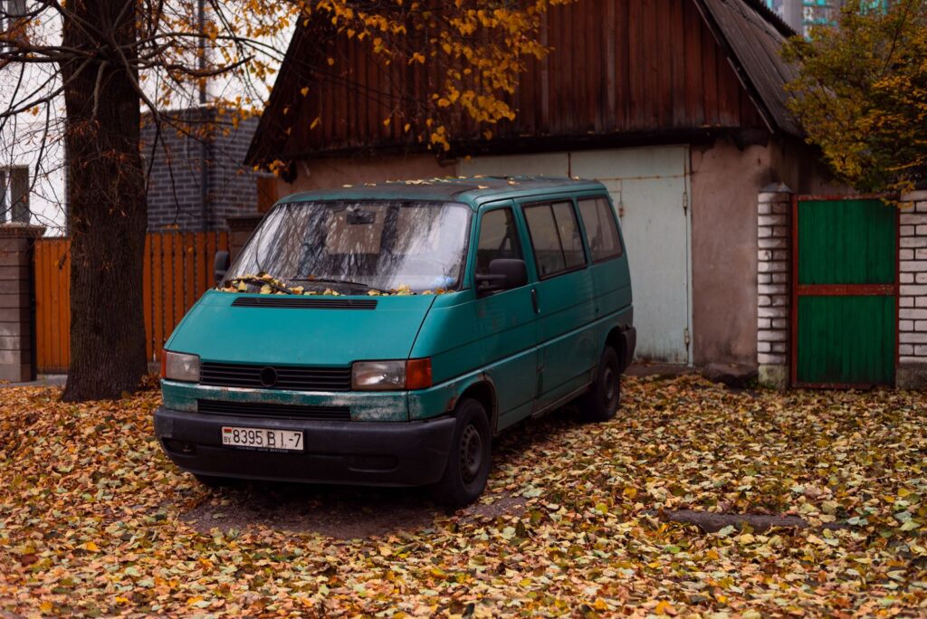 A vintage van surrounded by fallen autumn leaves, parked near a rustic wooden house with a green door.